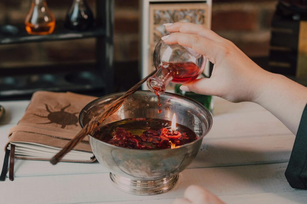 Close-up of a hand pouring red potion into a flame-lit bowl, suggesting a magical ritual.